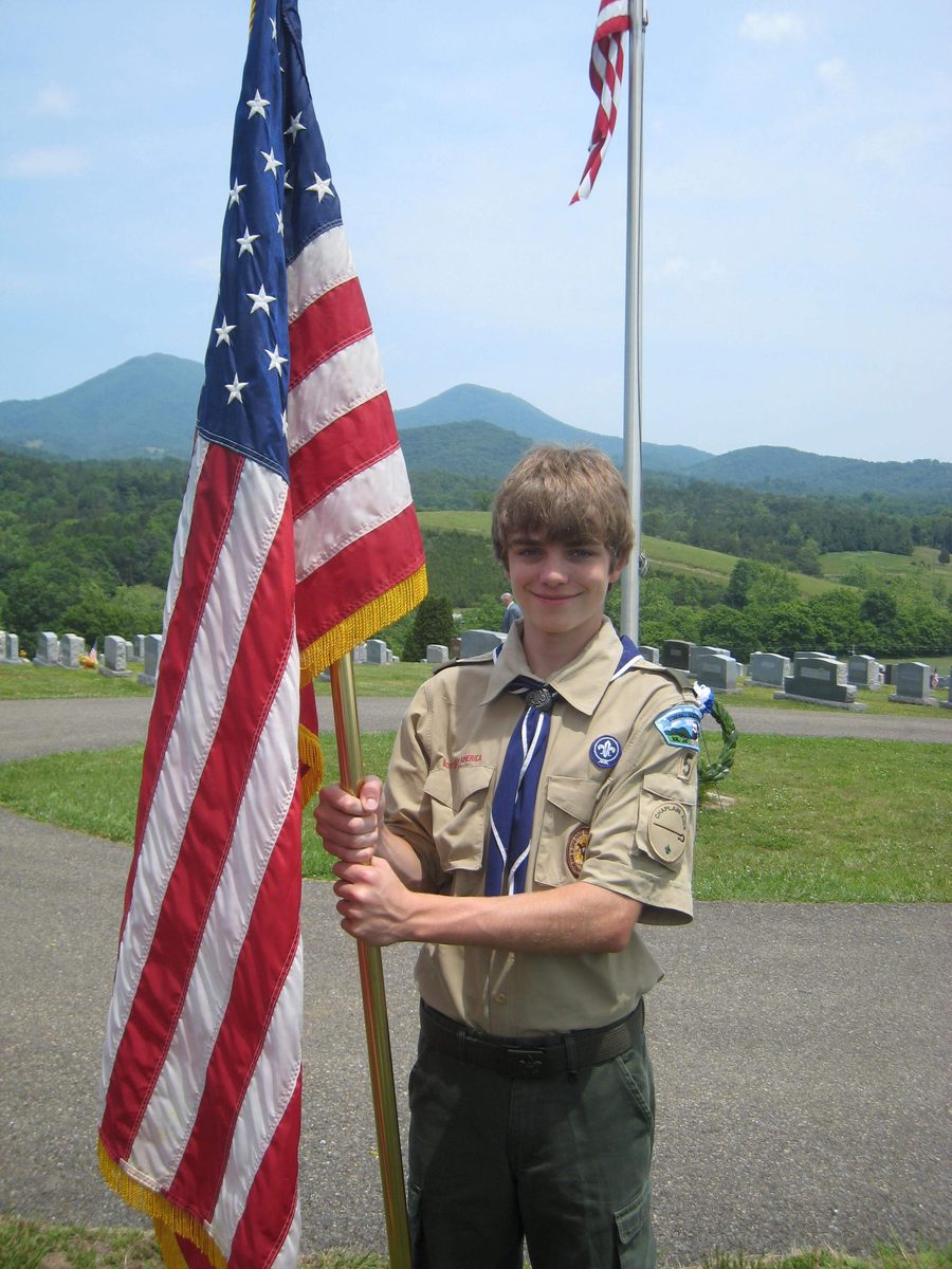 Ian as a teenager in his Boy Scout uniform and neckerchief, holding an American flag at a cemetery, with green hills and mountains behind him.