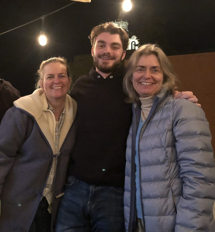 Ian outdoors at night under strings of light, standing between his mother Michelle and his aunt Stephanie with an arm around each of them, all three smiling.
