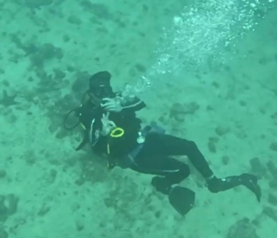 Ian scuba diving over a pale sandy seabed in clear green water, a trail of bubbles rising from his regulator.