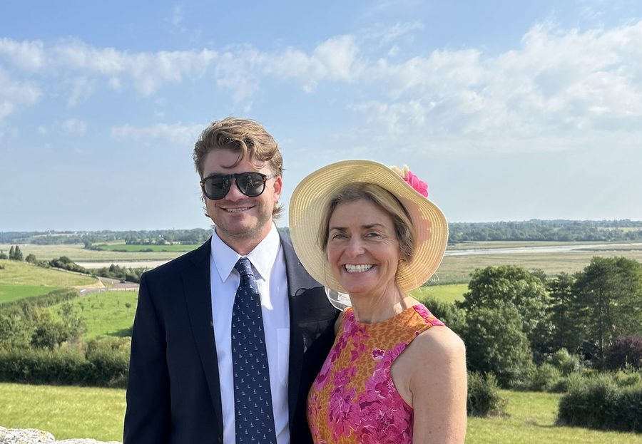 Ian in a dark suit and tie beside his mother Michelle in a wide straw hat and a pink and orange dress, green Normandy countryside and an estuary behind them.