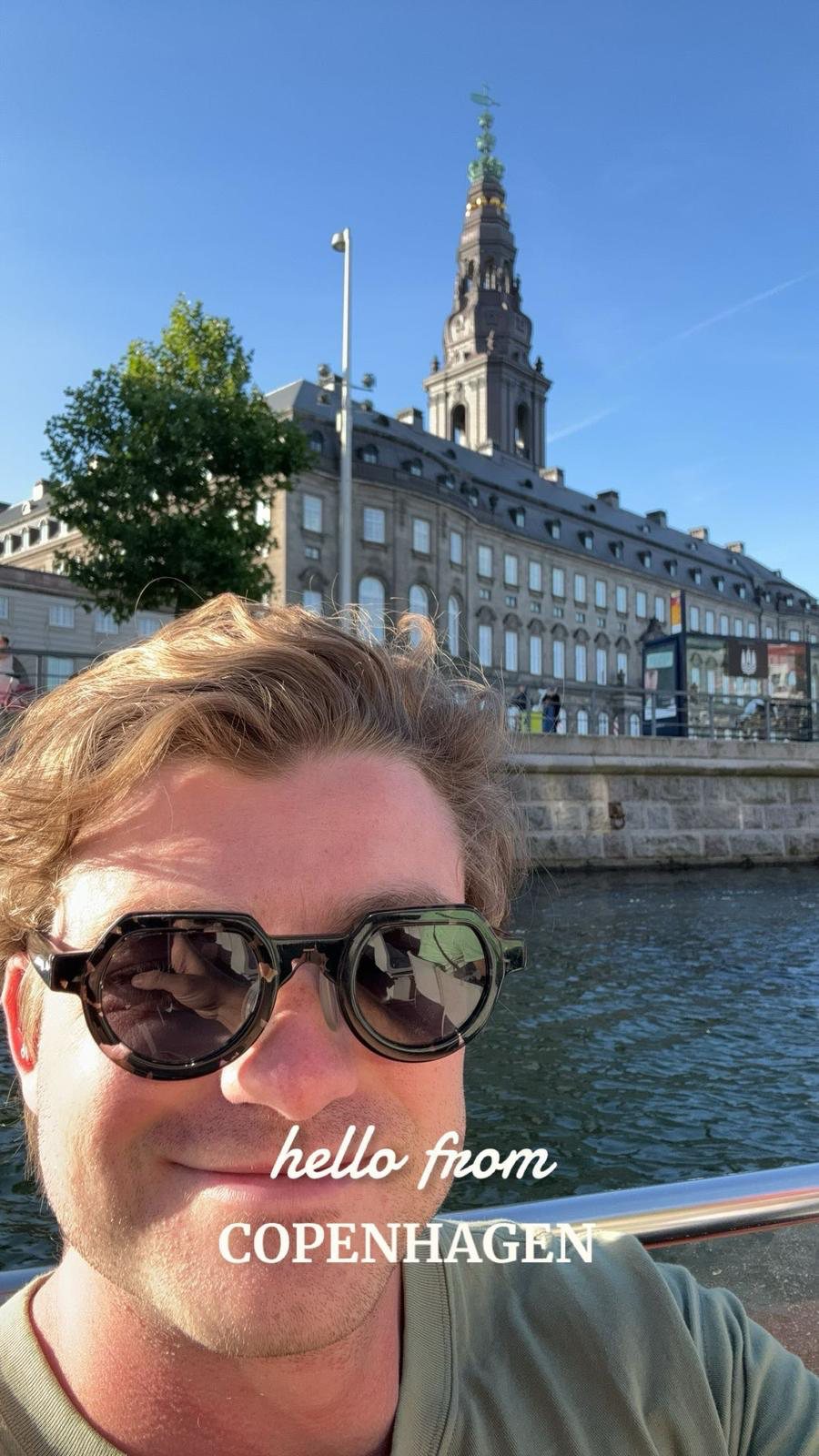 Ian in round sunglasses smiling into the camera from a boat on a Copenhagen canal, the long facade and green-topped spire of Christiansborg Palace behind him. Lettered across the photograph: hello from Copenhagen.