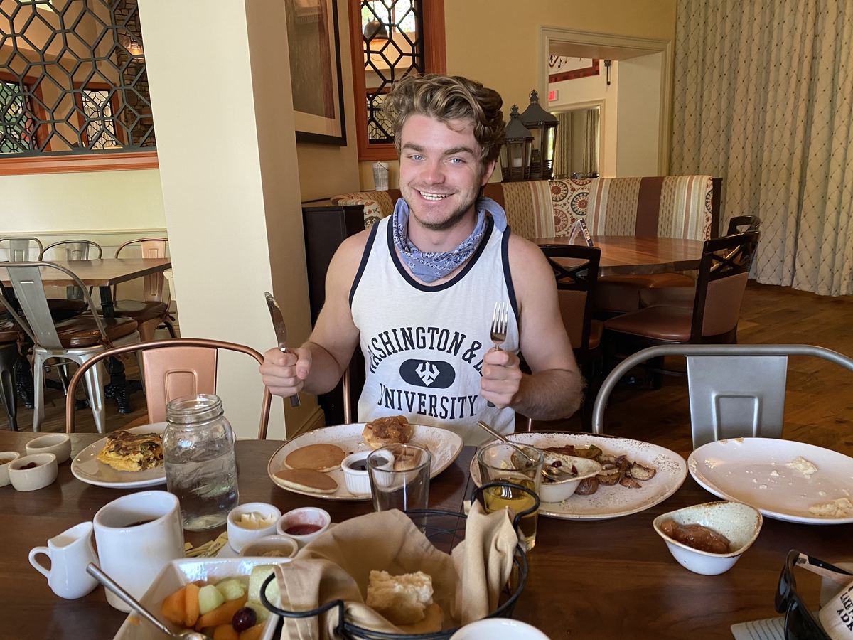 Ian grinning at a restaurant table with a knife in one hand and a fork in the other, wearing a Washington and Lee University tank top and a bandana, the table in front of him covered with pancakes, biscuits, potatoes, fruit and coffee.