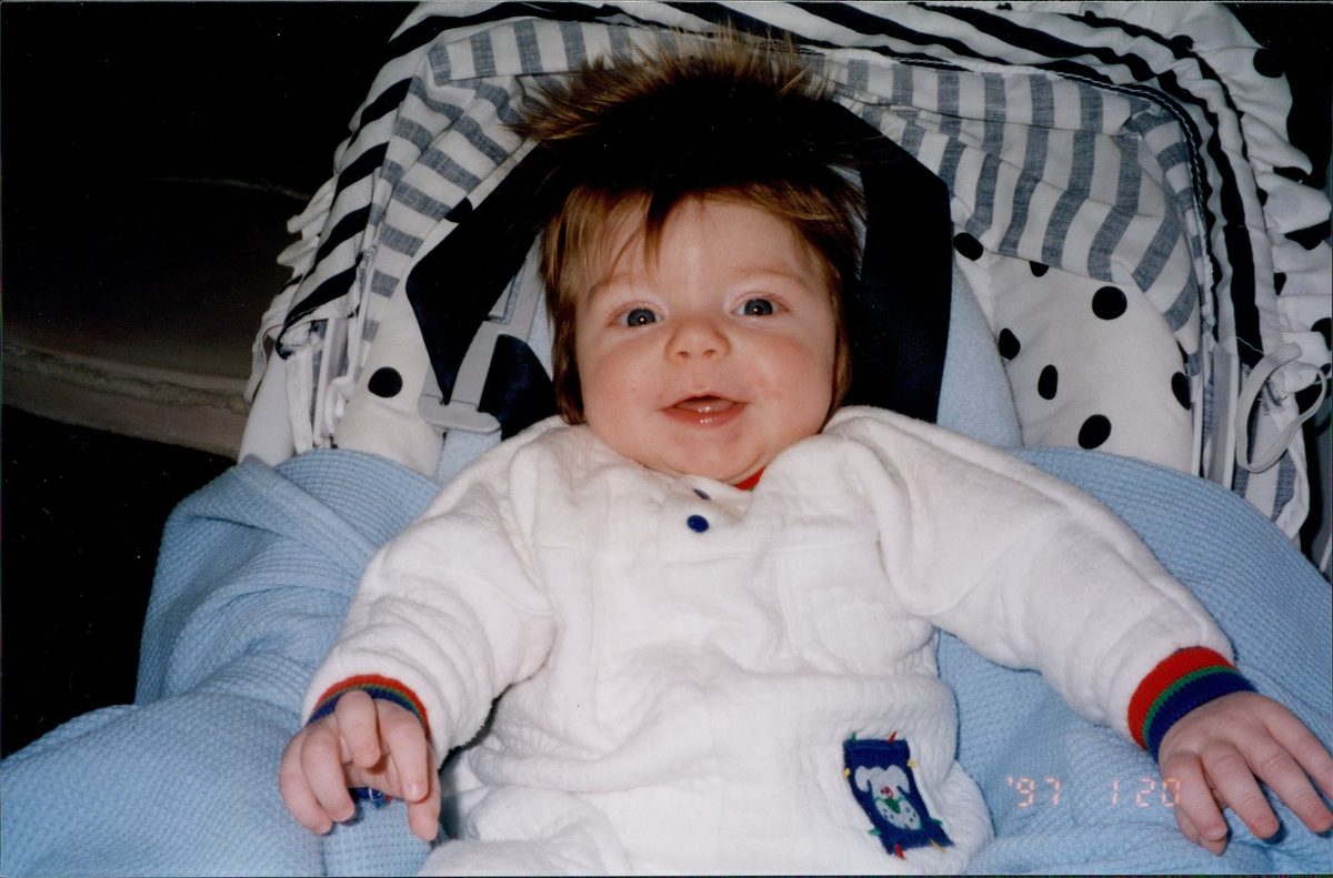 Ian as a baby, beaming up from a car seat under a striped canopy, in a white fleece suit on a pale blue blanket, his hair standing up. The film date stamp in the corner reads 97 1 20.