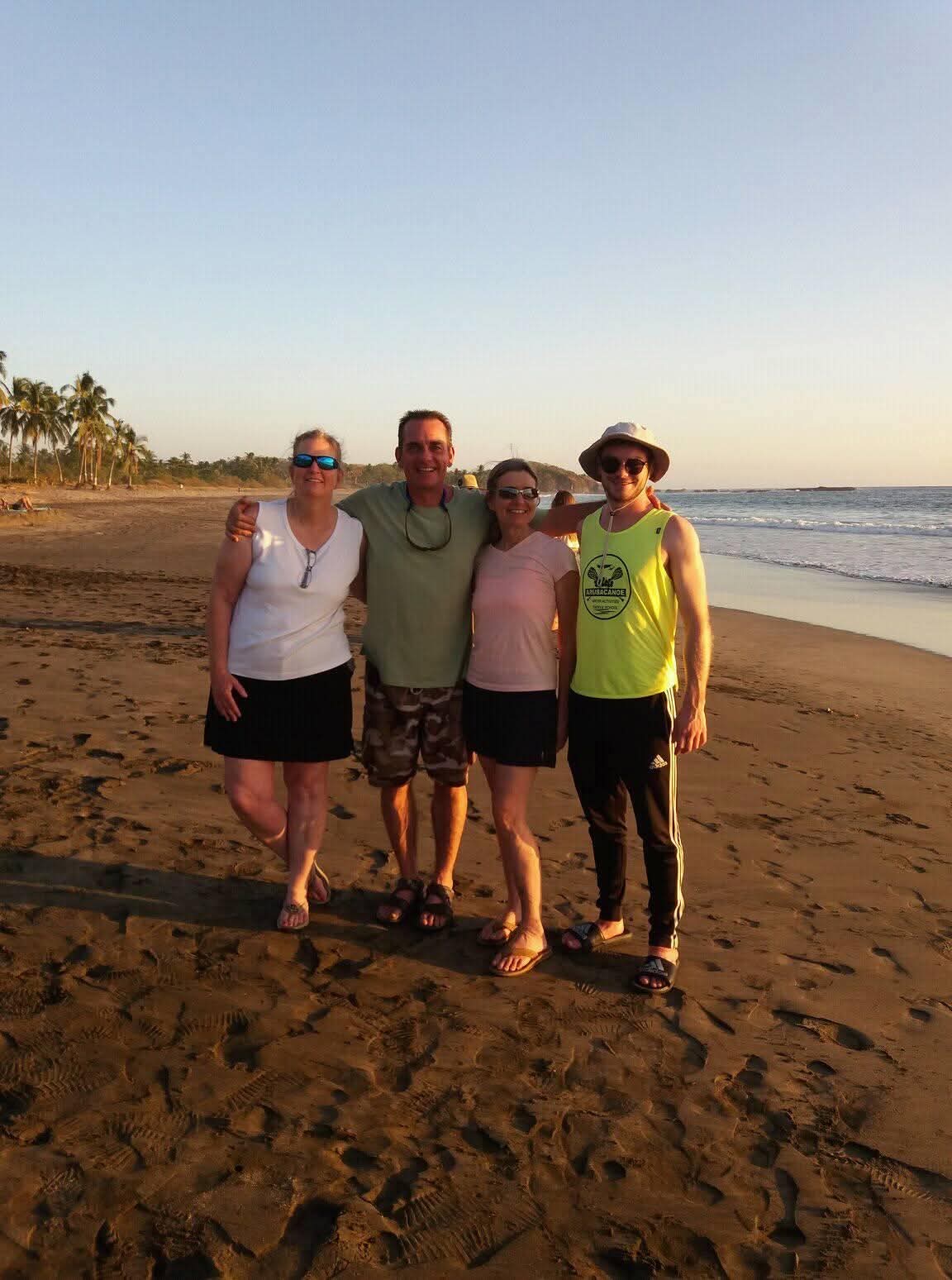 Ian with his mother, his uncle Scott and his aunt Stephanie, arms around one another on a beach at sunset, palm trees and the surf behind them.