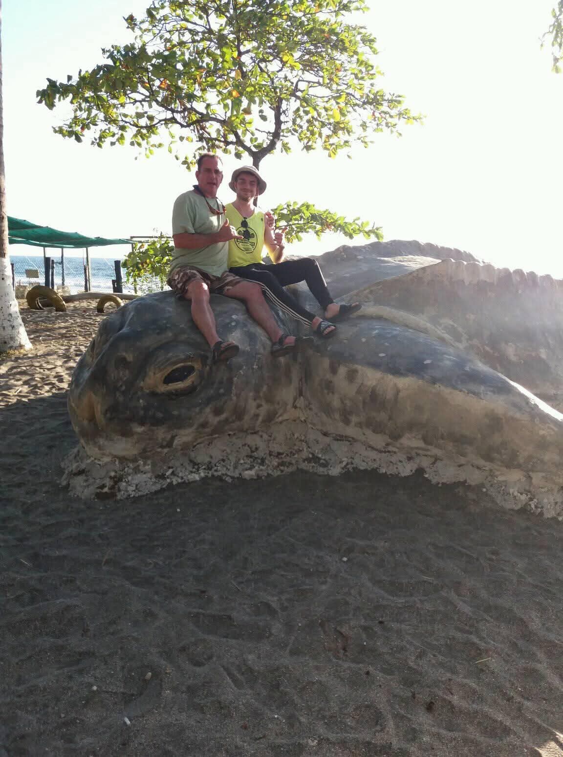 Ian and his uncle Scott sitting side by side on the head of a large sea turtle statue under a tree, both giving a shaka sign.