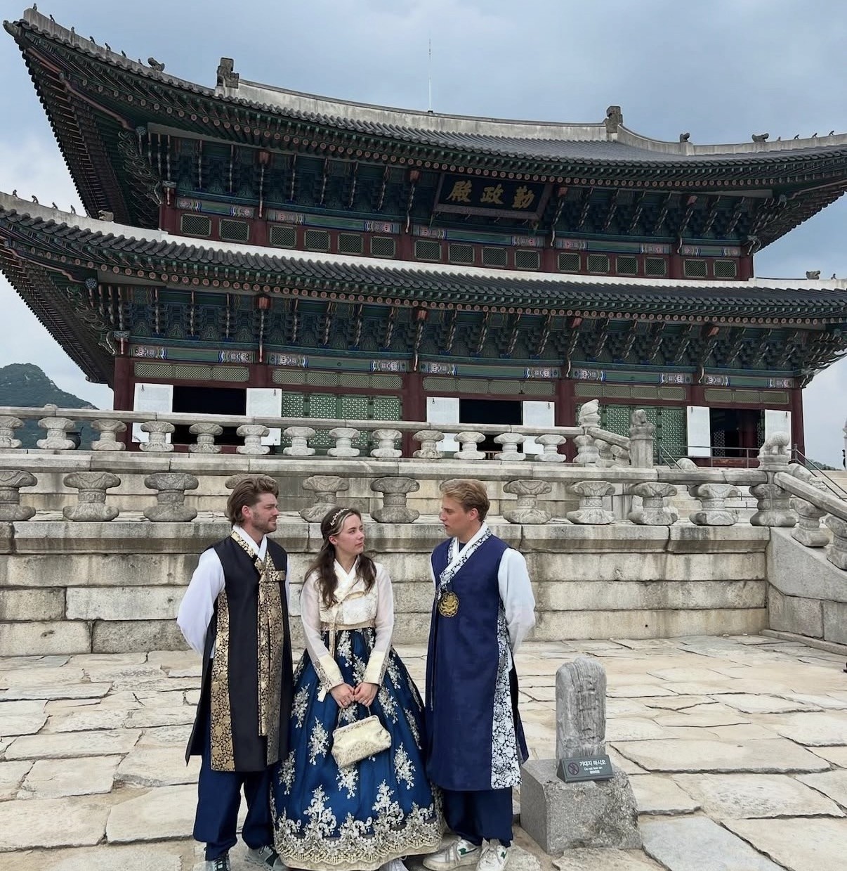 Ian and two friends dressed in traditional Korean hanbok, standing together in front of the ornate green-roofed Geunjeongjeon hall at Gyeongbokgung Palace, Seoul.