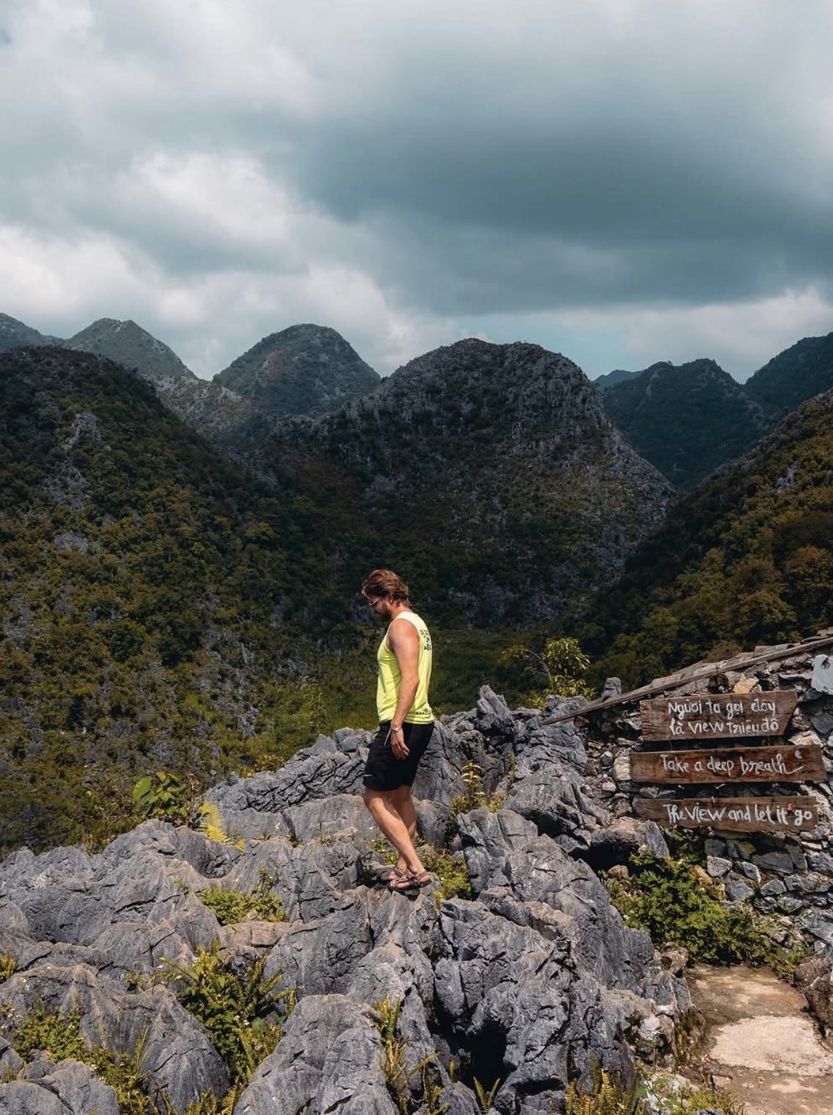 Ian, in a yellow tank top, standing on jagged limestone rock above a mist-covered mountain valley on the Ha Giang Loop, Vietnam, beside a wooden sign reading "Take a deep breath, the view and let it go."
