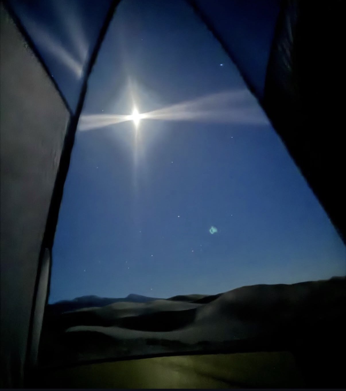 A bright moon with a cross-shaped flare, and a faint greenish glow among the stars, seen over a dark mountain ridge through the open door of a tent.
