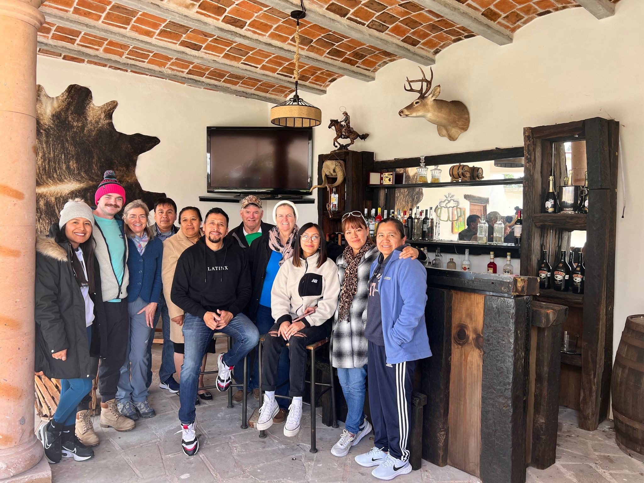 Ian, in a pink beanie and teal jacket, gathered with about a dozen family and friends for a Christmas photo in a rustic bar room in Mexico, a mounted deer head and cowhide on the wall behind them.