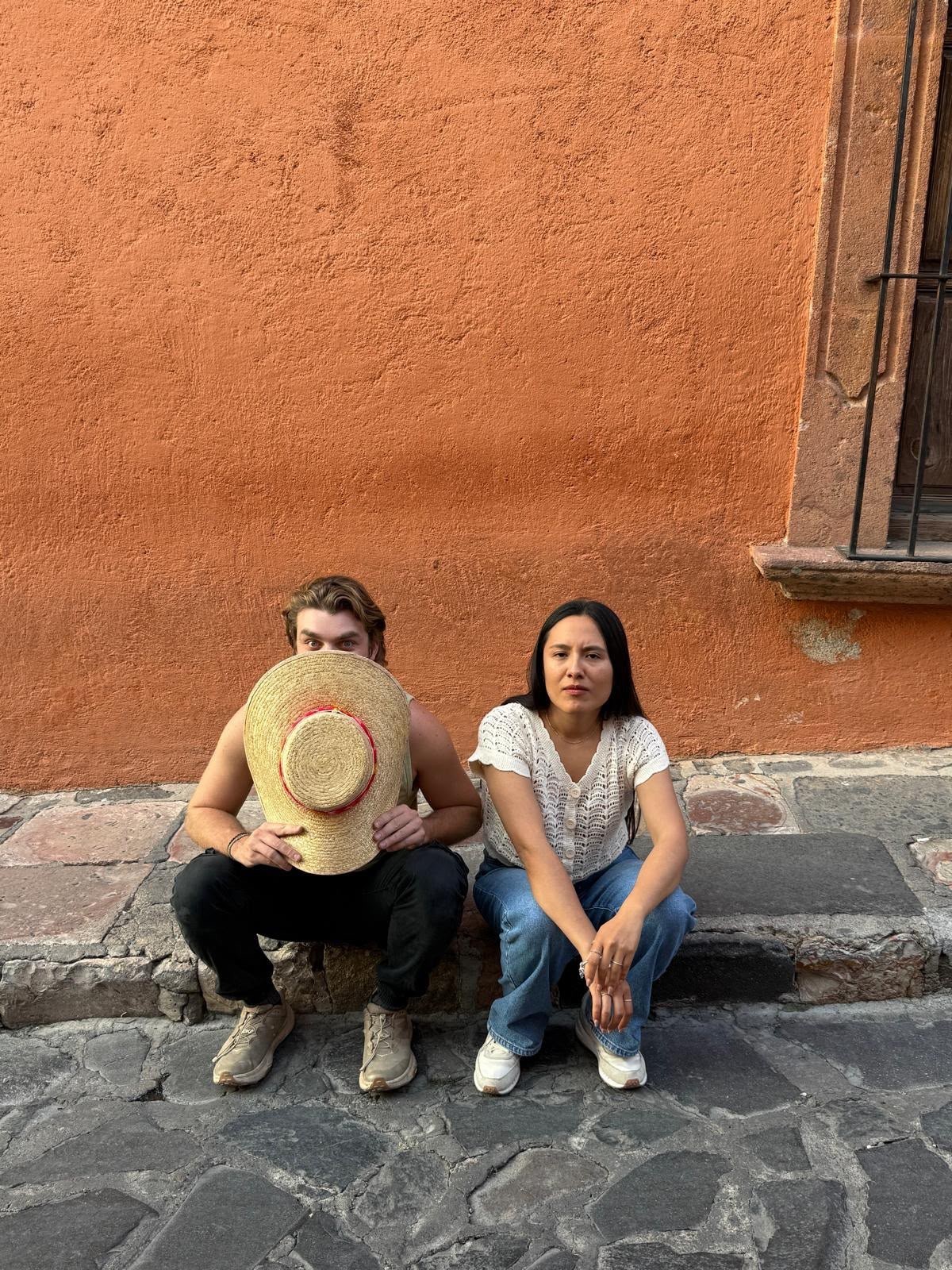 Ian, seated on a curb against a terracotta-orange wall, holding a wide-brimmed straw hat up in front of his face, beside his friend Jazmin.