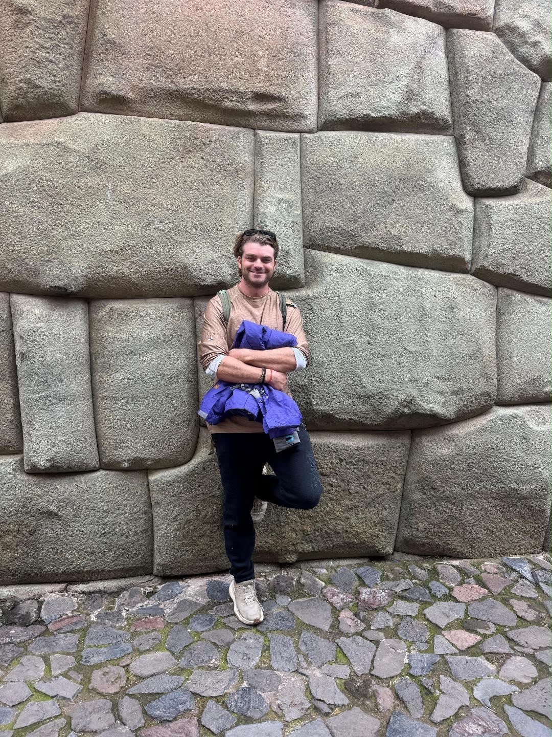 Ian smiling in front of a massive wall of precisely fitted Incan stone blocks, a folded jacket tucked under one arm.