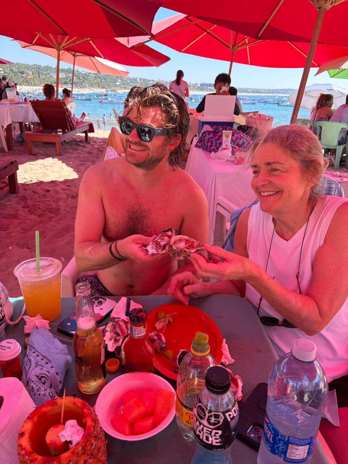 Ian, shirtless and sunglassed with wet hair, sharing fresh oysters with his mother Michelle at a table under red beach umbrellas, the ocean behind them.