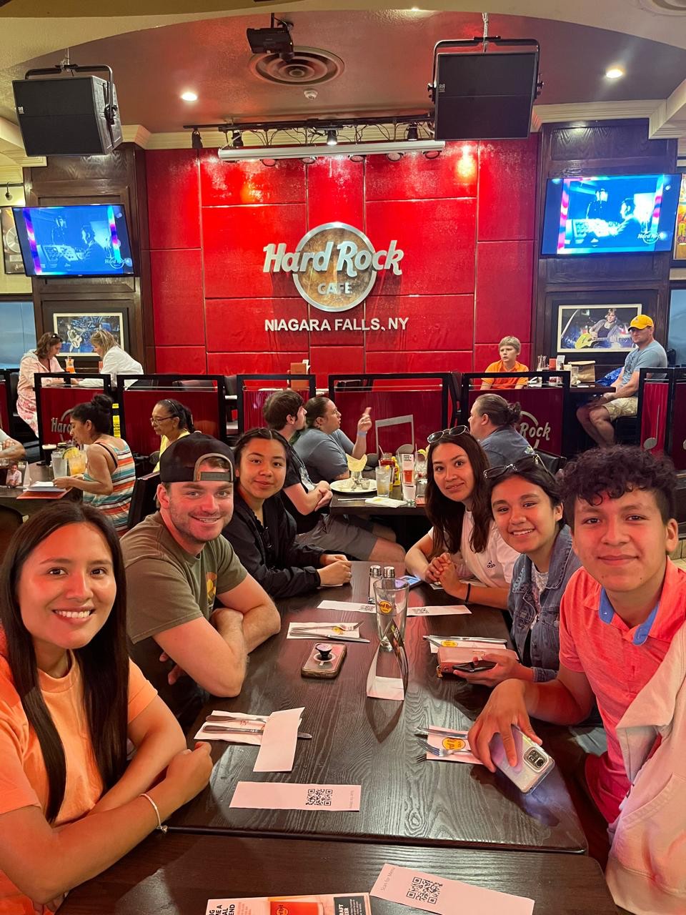 Ian, in a cap and olive shirt, gathered around a restaurant table with a group of friends beneath a lit "Hard Rock Cafe, Niagara Falls, NY" sign.