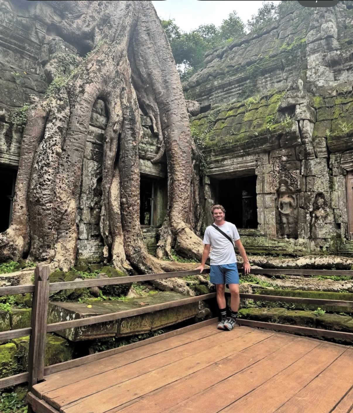 Ian standing on a wooden walkway at the Ta Prohm temple ruins in Cambodia, dwarfed by massive tree roots engulfing the ancient stone towers beside a carved stone figure.