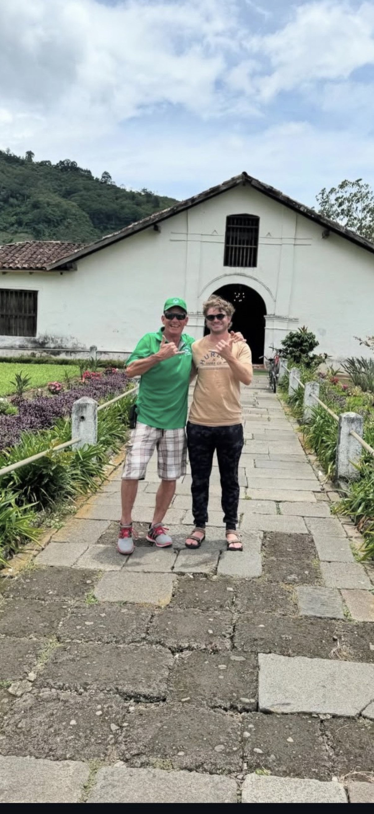 Ian and his uncle Scott, both giving a shaka sign, standing arm in arm on a stone path in front of a white colonial-style building with a tiled roof, green mountains behind.