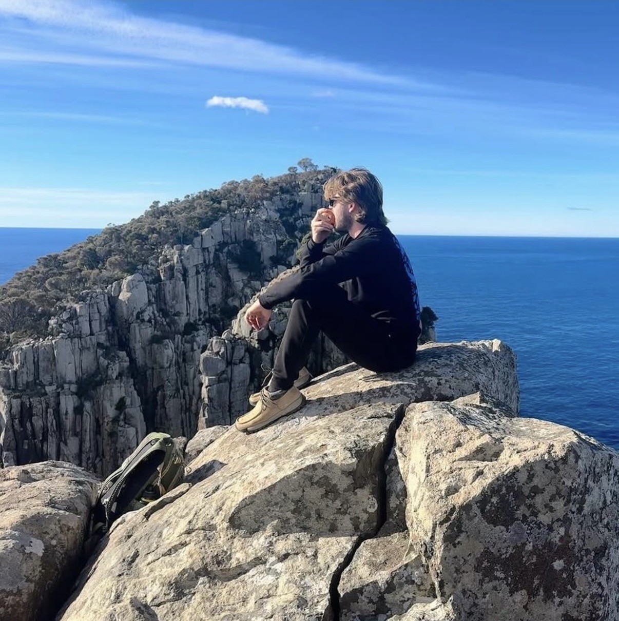 Ian sitting cross-legged on a rocky cliff edge overlooking the ocean and a mountain ridge in the Western Arthur Range, Tasmania, eating an apple.