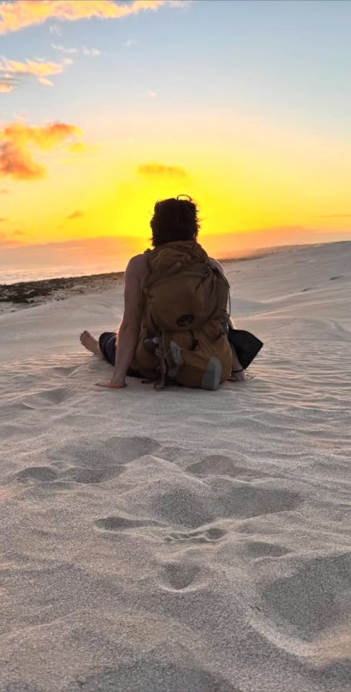 Ian, seen from behind and wearing a hiking backpack, sitting on a sand dune watching a vivid orange and yellow sunset over the ocean in Australia.