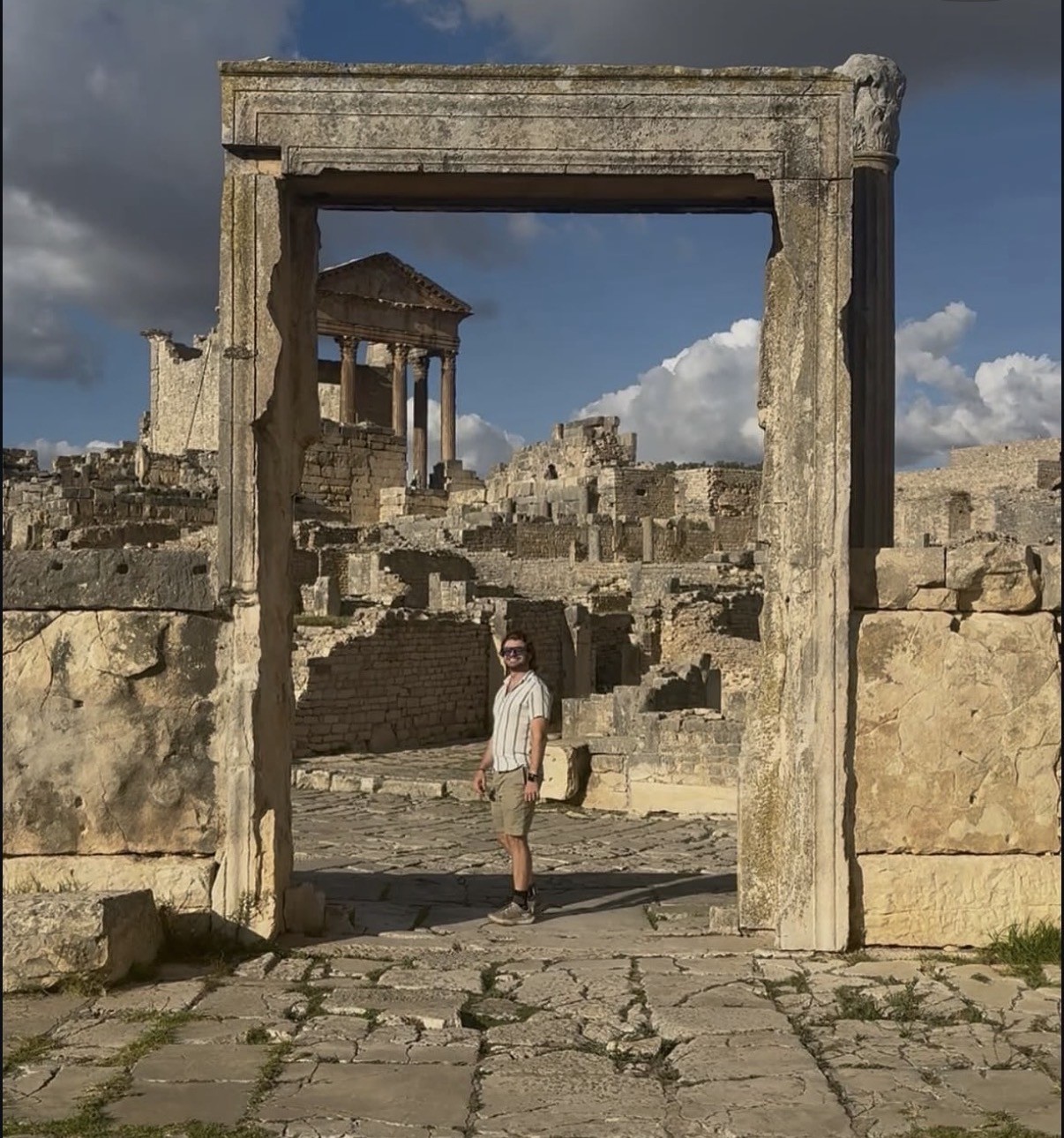Ian standing beneath a stone archway at the ancient Roman ruins of Dougga (Thugga) in Tunisia, a colonnaded temple visible in the background.