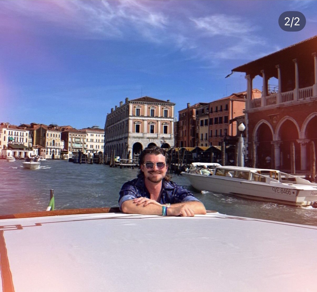Ian, in sunglasses, leaning on the rail of a boat on Venice's Grand Canal, historic waterfront buildings and other boats behind him.
