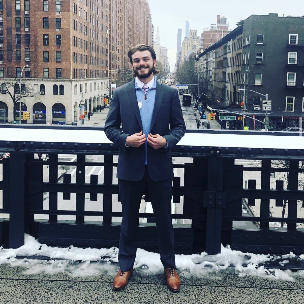 Ian, in a suit and tie, smiling on New York City's High Line with the Manhattan skyline and a snow-dusted railing behind him.