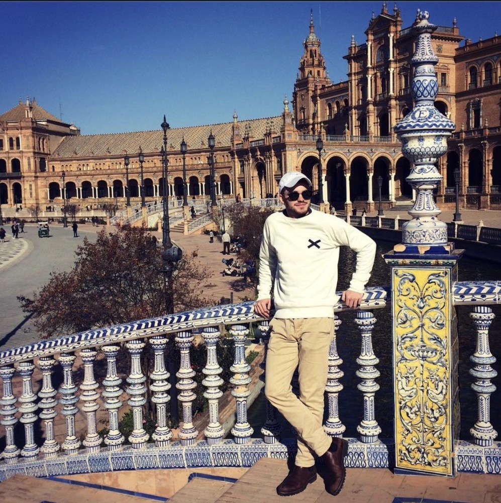 Ian, in a white cap and sunglasses, leaning against an ornately tiled balustrade above the moat at the Plaza de España in Seville, Spain.
