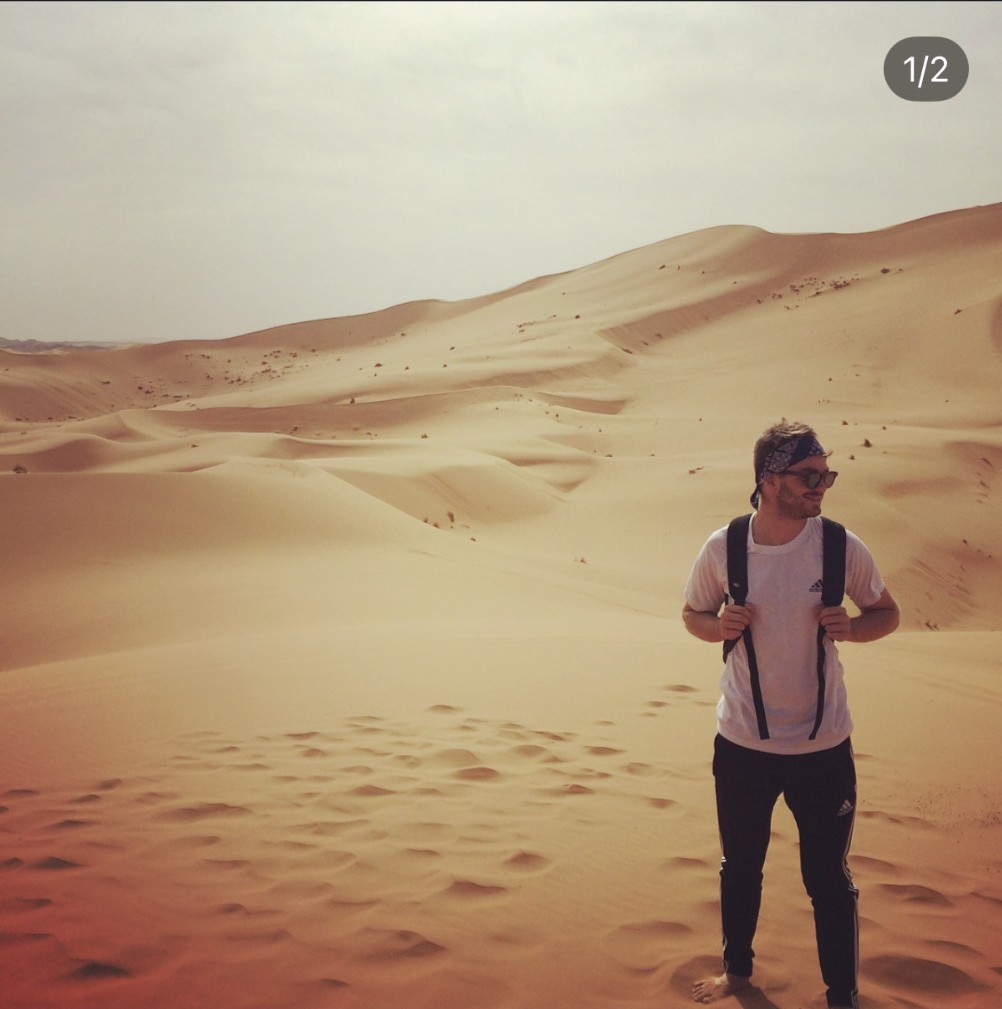 Ian, wearing a patterned bandana and a backpack, standing among the sand dunes of the Sahara Desert in Morocco.