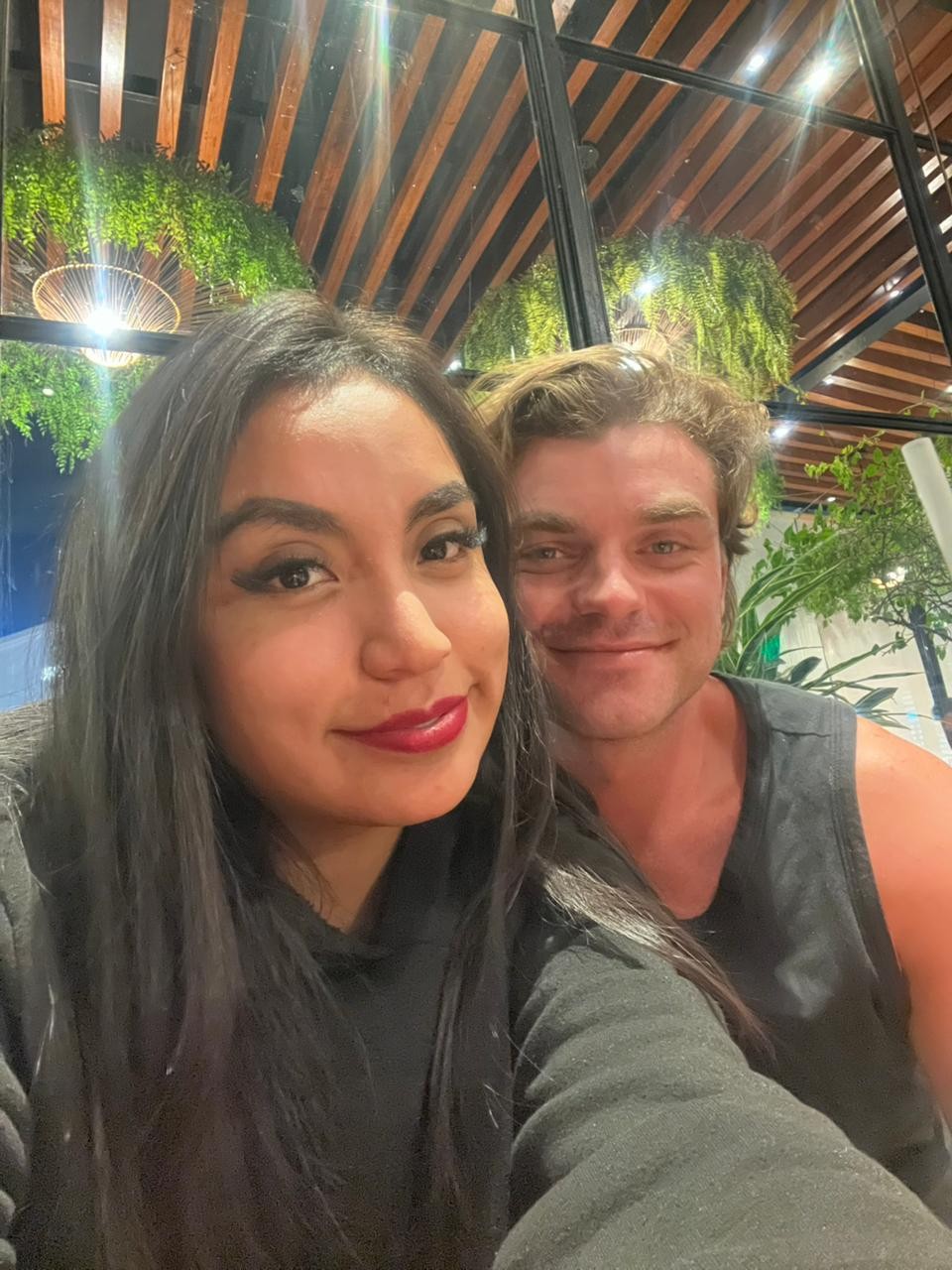 Ian and his friend Silvana taking a smiling selfie together beneath a plant-covered wooden ceiling.