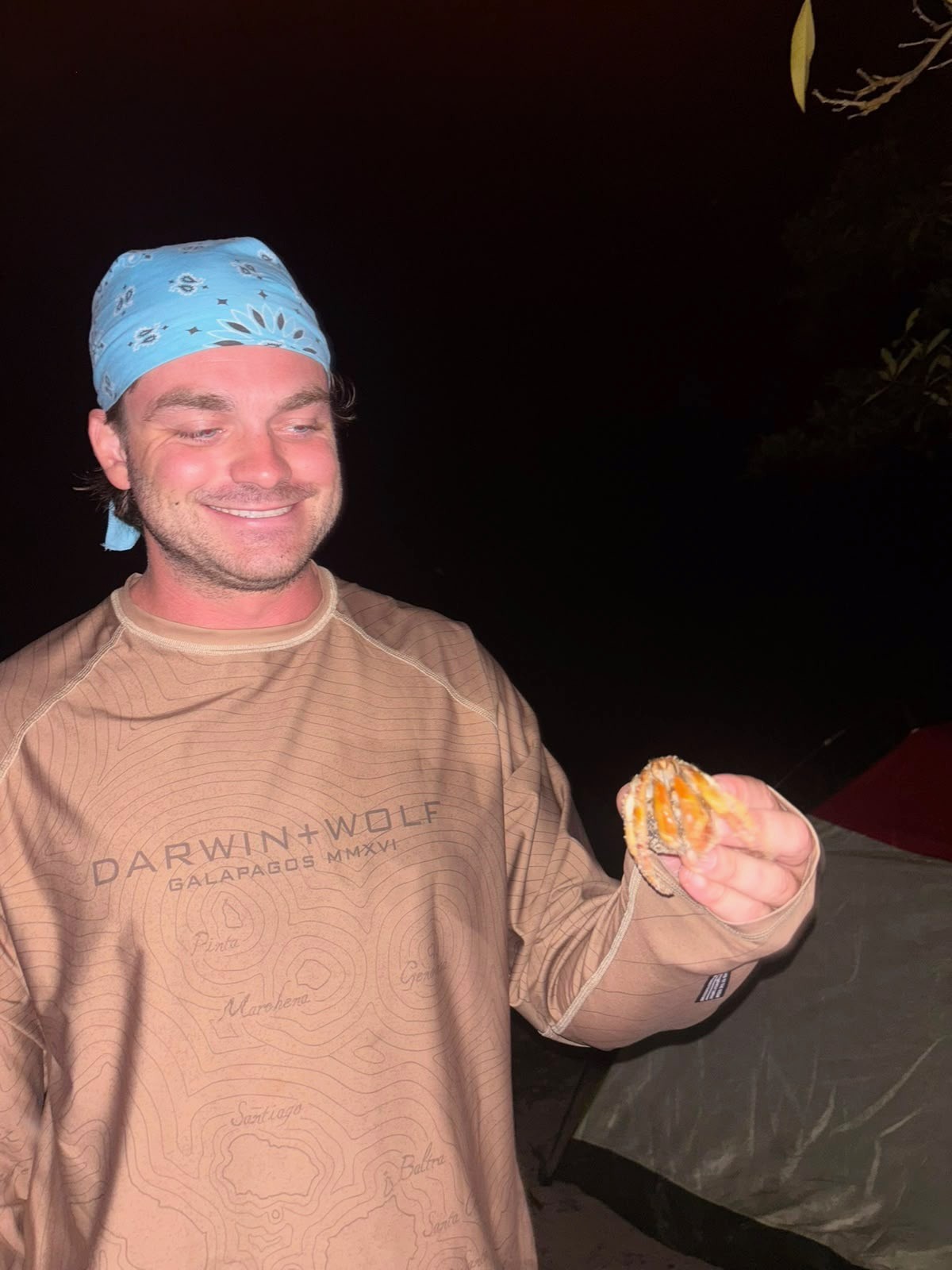 Ian, wearing a blue bandana and a shirt printed with a map of the Galápagos Islands, smiling at night while holding up a small crab to look at it.