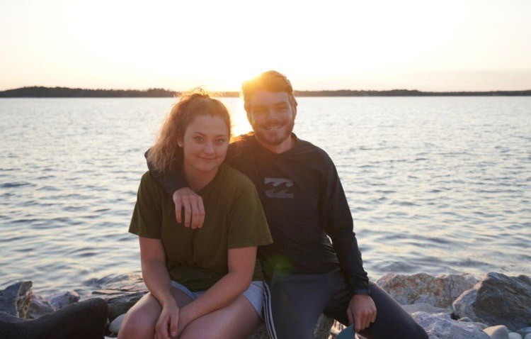 Ian and his friend Marisa sitting together on rocks at the water's edge in Nova Scotia at sunset, her hand resting on his shoulder.