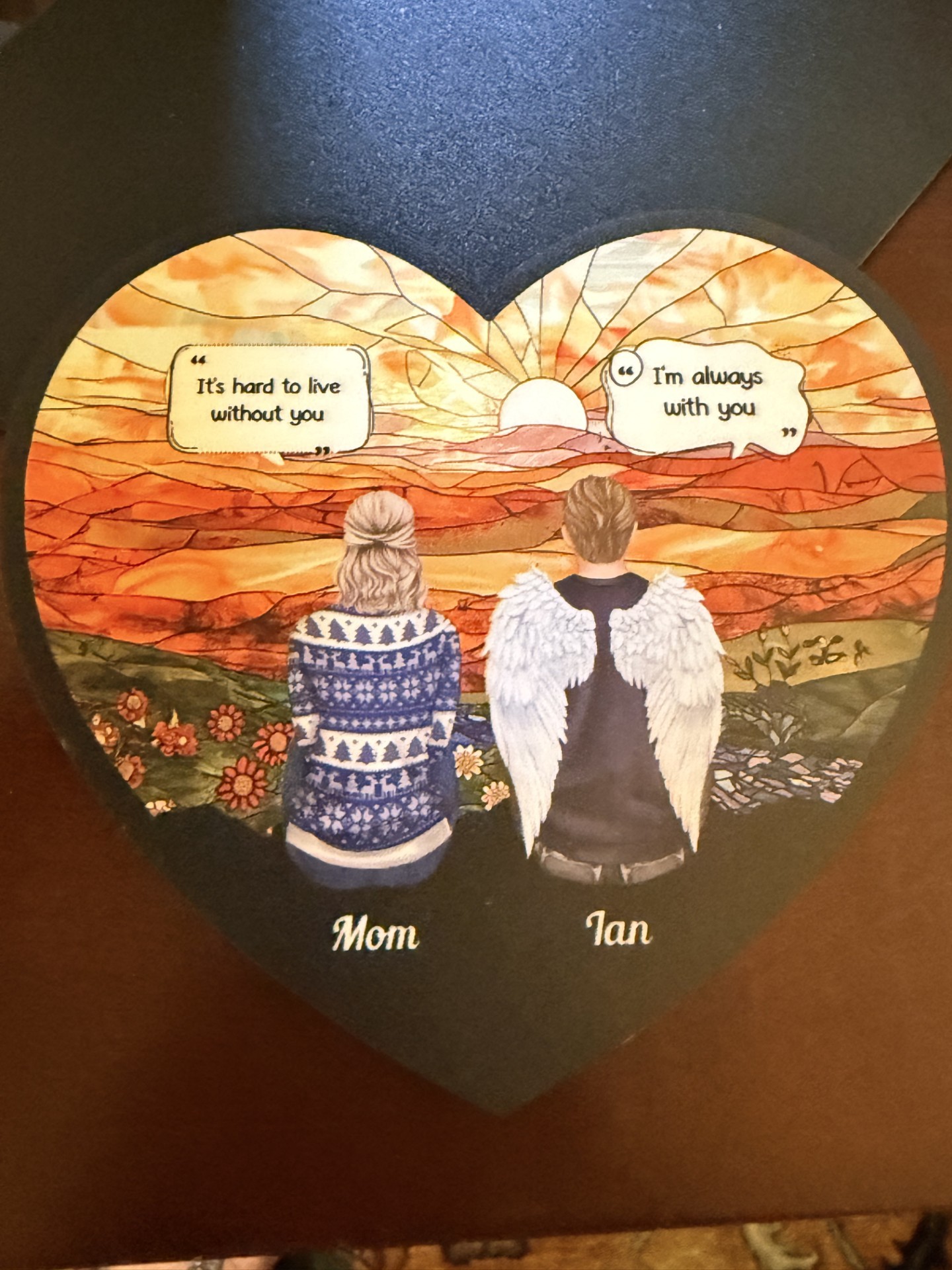 An illustrated memorial keepsake: Ian, with angel wings, and his mother sitting together watching a sunset, with the words "It's hard to live without you" and "I'm always with you" beside them.
