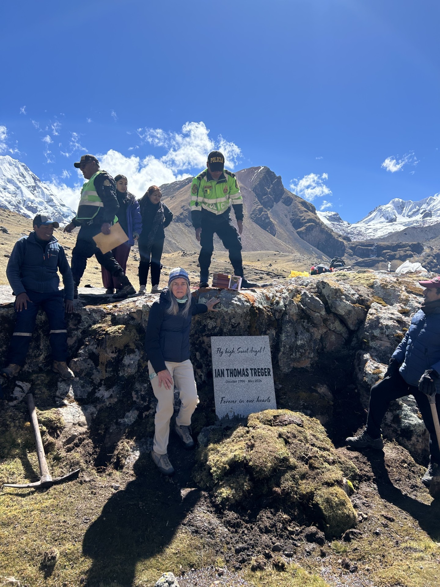Ian's mother Michelle beside a memorial stone reading "Fly high, Sweet Angel! Ian Thomas Treger, October 1996 &ndash; May 2026, Forever in our hearts," set among high rocks in the Peruvian Andes, surrounded by Peruvian police and friends who helped carry the stone with Ian's ashes to this spiritual place, snow-capped mountains behind.
