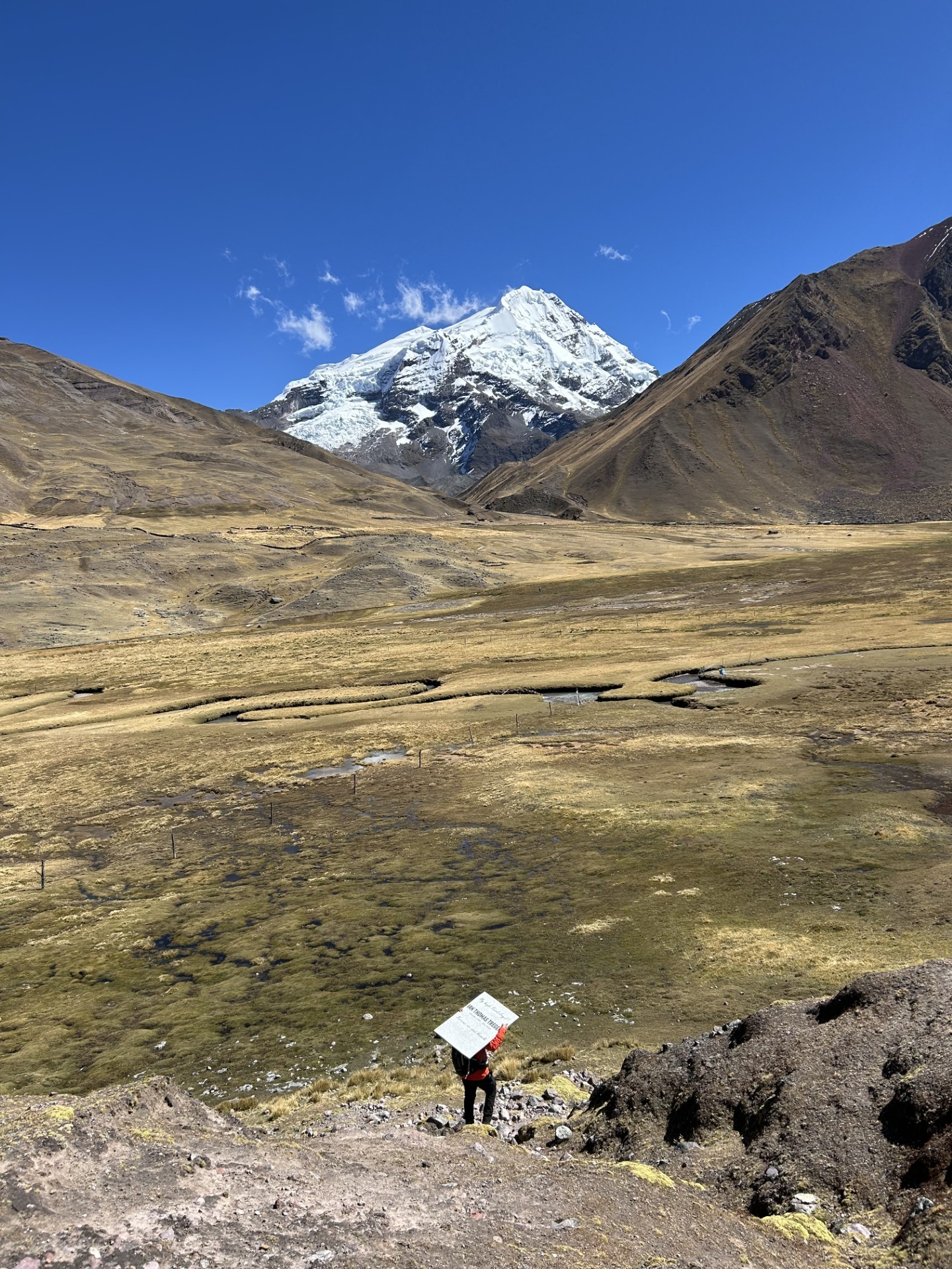 Henrry, seen from a distance, carrying Ian's memorial stone above his head across a vast Andean valley in Peru, a snow-capped peak in the background.