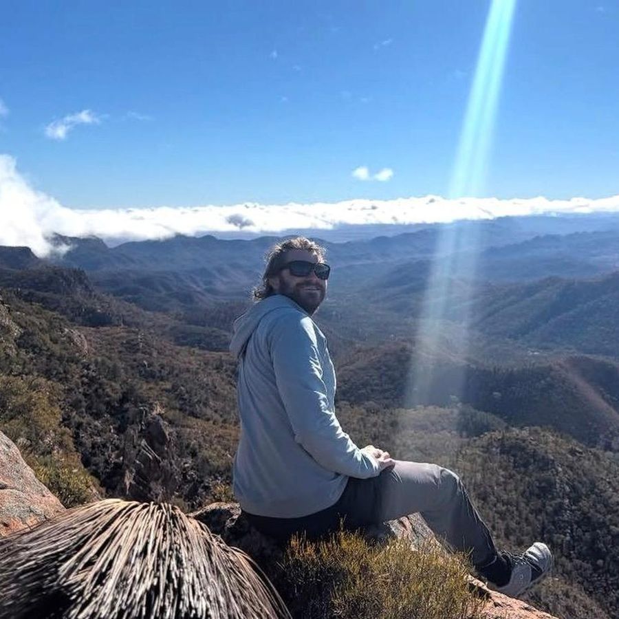 Ian Treger sitting on a rocky summit in sunglasses, looking back at the camera above a valley and a bank of cloud.