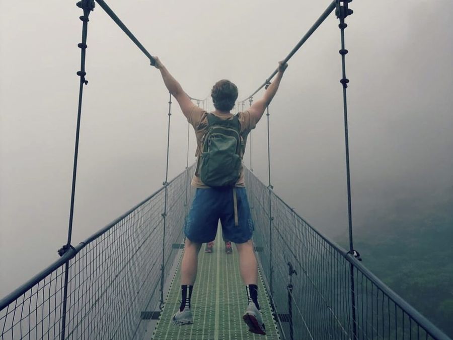 Ian Treger, back to the camera, gripping the cables of a fog-shrouded suspension bridge.